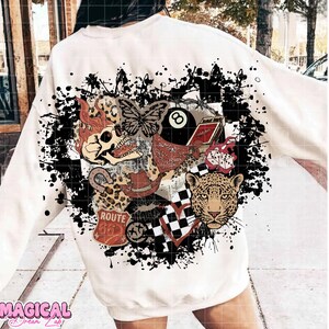 May include: Cream-colored sweatshirt with a graphic design featuring a skull, butterfly, leopard, and other vintage-style elements. The design includes a Route 66 sign, an 8-ball, and a checkered heart, all set against a black splatter background.