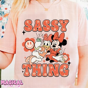 Puede incluir: Camiseta rosa claro con las palabras "SASSY THING" en naranja, con letras retro. La camiseta presenta a Daisy Duck tom&aacute;ndose una selfie con Minnie Mouse. Tambi&eacute;n se incluye un gr&aacute;fico de una flor sonriente.