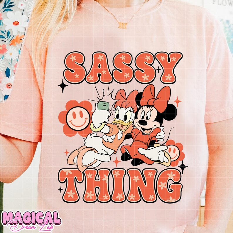 Puede incluir: Camiseta rosa claro con las palabras "SASSY THING" en naranja, con letras retro. La camiseta presenta a Daisy Duck tom&aacute;ndose una selfie con Minnie Mouse. Tambi&eacute;n se incluye un gr&aacute;fico de una flor sonriente.