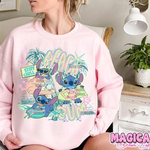 May include: Pink sweatshirt featuring a graphic of Stitch from Lilo & Stitch enjoying the beach. The design includes the words "BEACH SUM" and "STAY CHILL". Stitch is depicted surfing, drinking, and sunbathing. The words "ORIGINAL DESIGNER" are also present.
