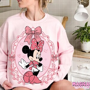 May include: A light pink sweatshirt featuring a Minnie Mouse graphic. The graphic includes Minnie Mouse in a pink dress and bow, framed by a pink oval with bows and hearts. The text "MAGICAL" is at the bottom right.