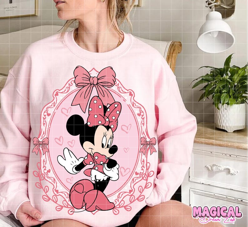 May include: A light pink sweatshirt featuring a Minnie Mouse graphic. The graphic includes Minnie Mouse in a pink dress and bow, framed by a pink oval with bows and hearts. The text "MAGICAL" is at the bottom right.
