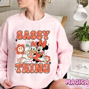Puede incluir: Sudadera rosa claro con la frase "SASSY THING" en fuente retro naranja. El dise&ntilde;o incluye a Daisy y Minnie Mouse haci&eacute;ndose un selfie. La sudadera tiene mangas largas y cuello redondo.