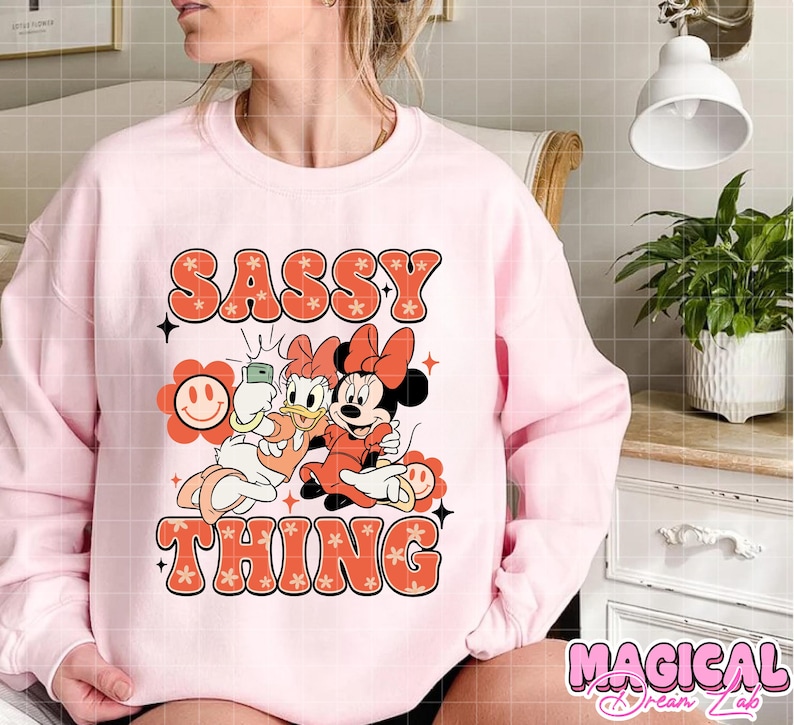 Puede incluir: Sudadera rosa claro con la frase "SASSY THING" en fuente retro naranja. El dise&ntilde;o incluye a Daisy y Minnie Mouse haci&eacute;ndose un selfie. La sudadera tiene mangas largas y cuello redondo.