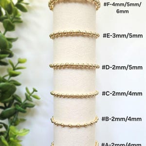 May include: Gold beaded rings displayed on a white cylinder. The rings vary in bead size and are labeled with sizes such as #F-4mm/5mm/6mm, #E-3mm/5mm, #D-2mm/5mm, #C-2mm/4mm, #B-2mm/4mm, and #A-2mm/4mm. Green foliage is visible on the left.