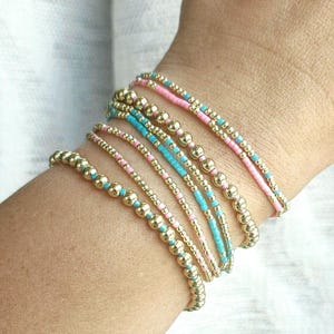 May include: A collection of beaded bracelets in gold, pink, and turquoise. The bracelets feature small gold beads interspersed with pink and turquoise seed beads. The bracelets are stacked on a wrist, showcasing the different color combinations.