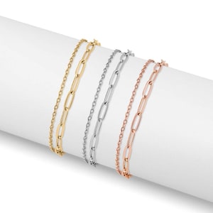 May include: Three delicate chain bracelets in gold, silver, and rose gold. Each bracelet features a different chain style, including a classic cable chain and a paperclip chain. The bracelets are displayed on a white surface.