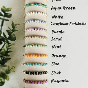 May include: A display of colorful beaded bracelets in various shades, including pink, aqua green, white, and purple. Each bracelet features gold-colored accents and is presented with corresponding color labels. The text includes "Pink", "Aqua Green", "White", "Cornflower Periwinkle", "Purple", "Sand", "Mint", "Orange", "Blue", "Black", "Magenta", and "Bright Pink".
