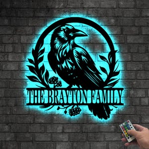 Könnte beinhalten: Schwarze Metall-Raben-Wandkunst mit türkisfarbenem Neonlicht. Der Rabe sitzt auf einem Ast, umgeben von einem kreisförmigen Rahmen und Blättern. Der Text "THE BRAYTON FAMILY" ist darunter zu sehen.