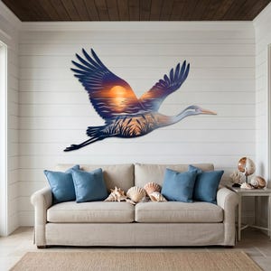 May include: A large metal wall art piece shaped like a flying crane, featuring a printed sunset scene over water with reeds. The bird is depicted in mid-flight with wings spread wide, showcasing a gradient of dark blue, orange, and yellow colors.