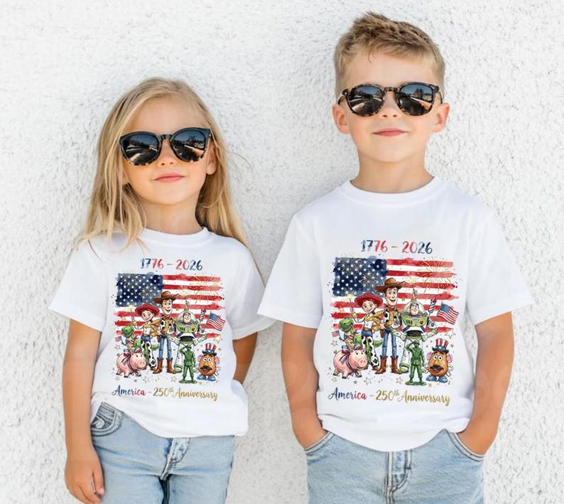 May include: Two white short-sleeve t-shirts featuring a graphic of Toy Story characters in front of a distressed American flag. The text reads 1776-2026 and America-250th Anniversary. The design includes Woody, Buzz Lightyear, Jessie, Rex, Hamm, and Mr. Potato Head.