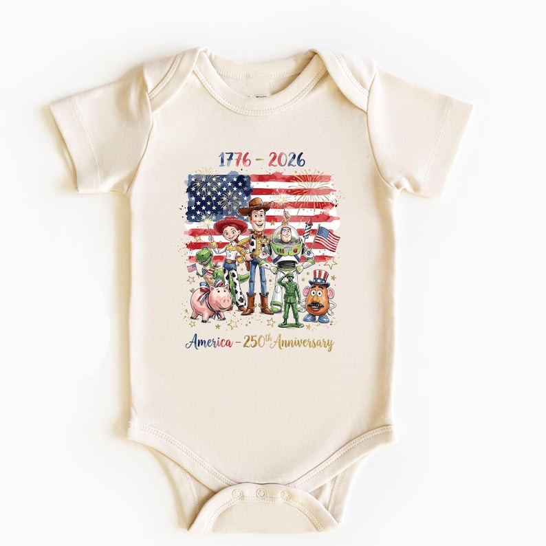 May include: A cream-colored baby bodysuit featuring a graphic print of Toy Story characters in front of an American flag. The text reads 1776-2026 and America-250th Anniversary. The design includes fireworks, stars, and patriotic colors.