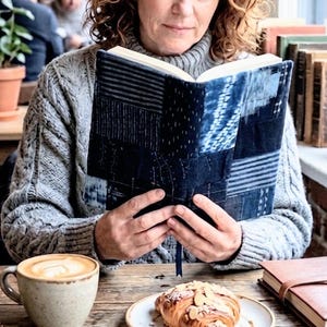 以下が含まれることがあります： 青と白のパッチワークの表紙の本を読む人。コーヒーカップ、ペストリー、革張りの本が木製のテーブルに置かれています。グレーのセーターが見えます。