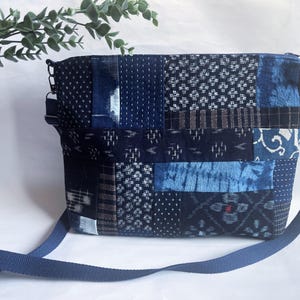 Może przedstawiać: Prostokątna torba na ramię z patchworkowym wzorem w odcieniach niebieskiego i białego. Torba posiada różne wzory tkanin, w tym kropki, paski i efekty tie-dye. Ma długi, regulowany niebieski pasek i zapięcie na zamek błyskawiczny.