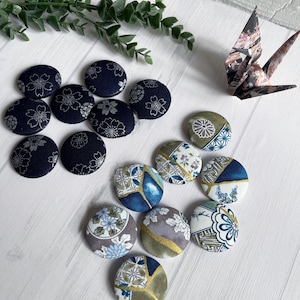 以下が含まれることがあります： 紺色、青色、金色、グレーの花柄や幾何学模様の布で覆われたボタンのコレクション。折り紙の鶴と緑も写っています。ボタンは丸く、手芸や縫製プロジェクトに適しています。