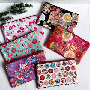 以下が含まれることがあります： 花柄の7つの長方形のジッパーポーチのコレクション。ポーチは、ピンク、青、紫、黒など様々な色で、赤または茶色のジッパーが付いています。デザインには、桜やその他の様式化された花が含まれています。