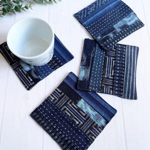 以下が含まれることがあります： 藍色の生地で作られたコースターのセット。ストライプやドットなど、さまざまな模様があります。白いセラミックカップがコースターの上に置かれています。コースターは白い木製の表面に配置されています。