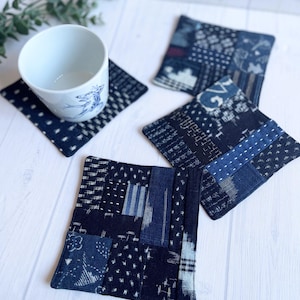 以下が含まれることがあります： 青い模様の白いセラミックカップが、ネイビーブルーのパッチワークコースターの上に置かれています。さまざまな模様のネイビーブルーのパッチワークコースターが3つあります。コースターは正方形で、布製です。