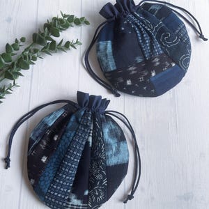 May include: Two round drawstring pouches with a patchwork design in shades of blue and navy. The pouches have a gathered top and a dark blue drawstring closure. The fabric features various patterns, including geometric and wave-like designs. The pouches are on a white wooden surface.