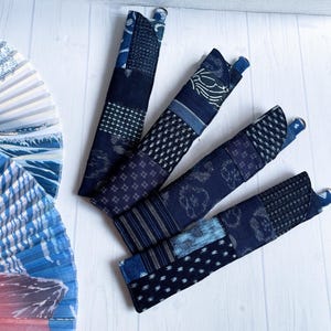 May include: Four long, narrow fabric pouches made from various shades of dark blue and indigo patchwork textiles. Each pouch features a small metal D-ring at the top and displays a variety of geometric and floral patterns in white and light blue.