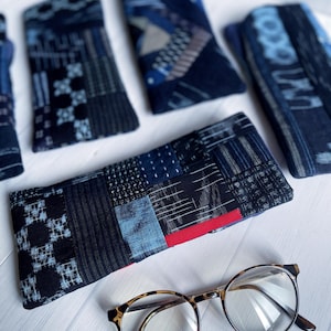 May include: A collection of rectangular fabric cases featuring various blue and black patchwork patterns with visible stitching, accompanied by a pair of round tortoiseshell-patterned eyeglasses positioned in the foreground.