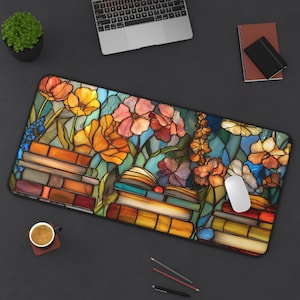 May include: A rectangular desk mat featuring a stained glass style illustration of stacked books and blooming flowers in shades of orange, yellow, pink, and blue. The design covers the entire surface of the mat, which is positioned on a dark desk.