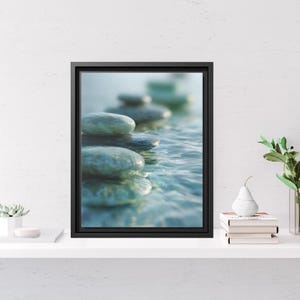 May include: A framed wall art print featuring a close-up photograph of smooth, rounded blue-grey stones stacked partially submerged in clear, rippling water. The artwork is enclosed in a thin, black rectangular frame with a deep profile.