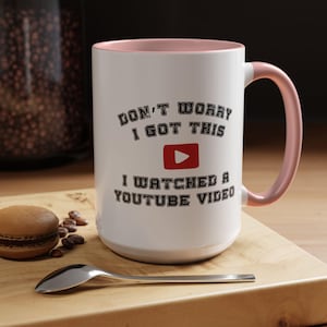 May include: White ceramic mug with a pink interior and handle. The mug features the text "DON'T WORRY I GOT THIS" above a red YouTube play button, and "I WATCHED A YOUTUBE VIDEO" below. A spoon and a macaron are on the wooden surface.