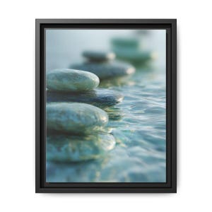 May include: A framed canvas print featuring a stack of smooth, gray-green stones partially submerged in rippling water. The stones are in focus, with a blurred background of more stones. The water has a blue-green hue.