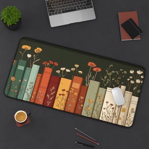 May include: A rectangular desk mat featuring a design of colorful books with flowers growing from the tops. The books are in shades of green, orange, and yellow. A white mouse and a cup of coffee are on the mat.
