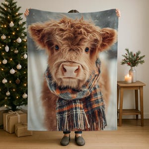May include: A cozy blanket featuring a Highland cow with long, shaggy, reddish-brown fur and blue eyes. The cow is wearing a plaid scarf with orange, blue, and brown tones. The background is a snowy scene.