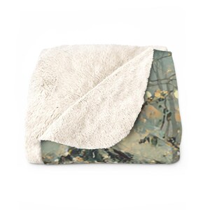 May include: A folded, cream-colored sherpa blanket with a nature-themed print. The print features a muted color palette of greens, browns, and yellows, suggesting a forest or landscape scene. The blanket's soft texture is visible.