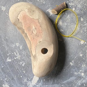 Puede incluir: Un objeto de madera de color claro, posiblemente una horma de zapato, con un agujero y una sección de veta de madera rojiza. También hay un trozo pequeño de madera y una cuerda amarilla.