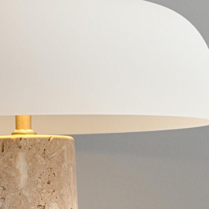 May include: A beige table lamp with a white lampshade. The lamp base is made of a textured, light-colored stone. The lampshade is a curved, off-white shape, casting a soft glow.
