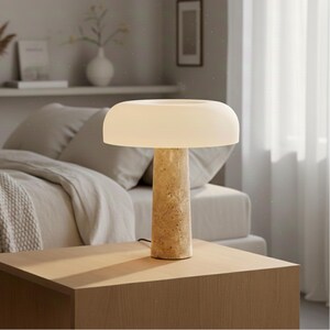 May include: A modern table lamp with a mushroom-shaped, off-white shade. The lamp has a cylindrical, textured base in a light beige color. The lamp is placed on a wooden nightstand.