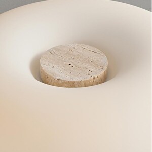 May include: A beige, circular stone bottle stopper sits in a white, concave surface. The stone stopper has a textured surface with small holes. The background is a soft, off-white color.