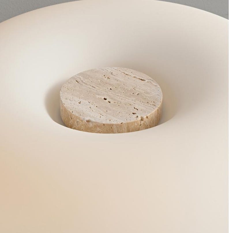 May include: A beige, circular stone bottle stopper sits in a white, concave surface. The stone stopper has a textured surface with small holes. The background is a soft, off-white color.