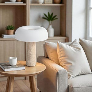May include: A modern table lamp with a white, mushroom-shaped shade and a beige, textured stone base. The lamp sits on a round, wooden side table next to a small white cup and a book. The background includes a sofa and a wooden shelf.