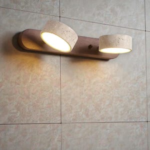 Peut inclure: Un luminaire mural moderne avec deux lampes cylindriques. Les lampes sont faites d'un matériau texturé de couleur claire et sont montées sur une base en bois. Une lampe est inclinée, et les deux émettent une lueur chaleureuse. Pour l'intérieur.