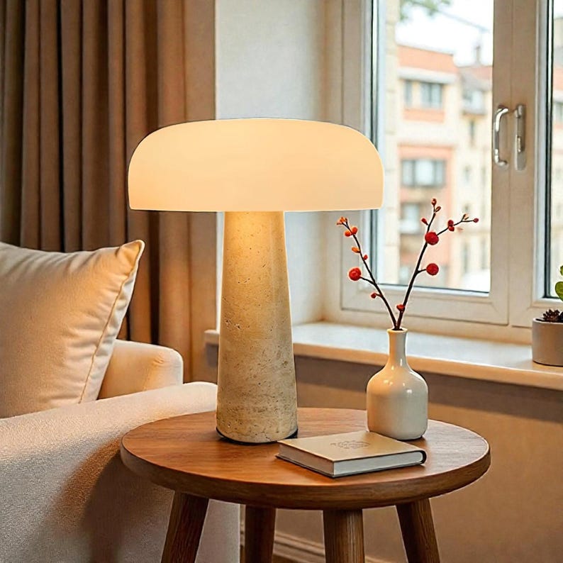 May include: A modern table lamp with a cream-colored, rounded shade and a textured, beige stone base. The lamp sits on a small, round wooden table. A small white vase with red berries and a closed book are also on the table.