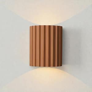 May include: A brown, cylindrical wall sconce with a ribbed design. The sconce is mounted on a light gray wall, and the light shines up and down, creating a soft glow. The fixture is a modern design.