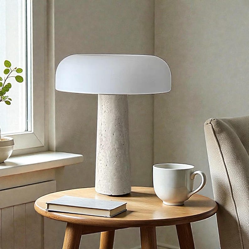 May include: A modern table lamp with a white, rounded shade and a textured, cylindrical base. The lamp sits on a wooden side table next to a white mug and a closed book. The lamp's design is minimalist.