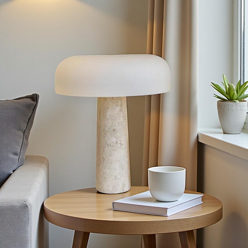 May include: A modern table lamp with a cream-colored, rounded shade and a textured, beige stone base. The lamp sits on a round wooden table, next to a white cup and a book. A small potted plant is visible in the background.