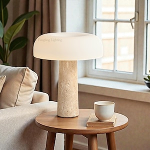 May include: A modern table lamp with a cream-colored, mushroom-shaped shade and a textured, beige stone base. The lamp sits on a round wooden side table, next to a small white cup and a book. The lamp is labeled "Fangfang Lighting".