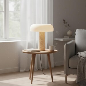 May include: A modern table lamp with a cream-colored shade and a textured, cylindrical base. The lamp sits on a small, round wooden side table, alongside a book and a mug. The scene is set in a well-lit living room.
