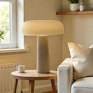May include: A modern table lamp with a beige, mushroom-shaped shade and a textured, cylindrical base. The lamp sits on a round wooden side table. A white mug is on the table. The lamp provides soft, ambient lighting, suitable for a living room or bedroom.