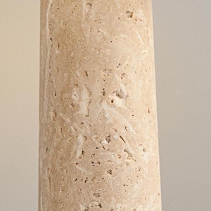 May include: Close-up of a cylindrical, beige stone column with a rough, textured surface. The stone has a natural, porous appearance with small holes and variations in color, creating a unique, organic aesthetic. The column is likely a decorative element.