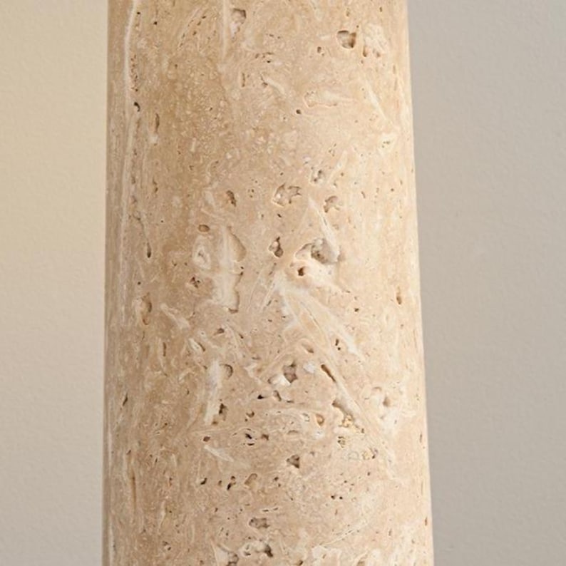 May include: Close-up of a cylindrical, beige stone column with a rough, textured surface. The stone has a natural, porous appearance with small holes and variations in color, creating a unique, organic aesthetic. The column is likely a decorative element.