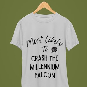Puede incluir: Camiseta gris claro con el texto negro "Most Likely To Crash The Millennium Falcon". La camiseta está colgada de una percha de madera sobre un fondo verde.