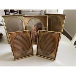 May include: A set of five rectangular gold-toned picture frames with oval-shaped inner matting. Three frames feature empty oval openings, while two frames contain ribbed brown inserts. One frame displays a photograph of a person.